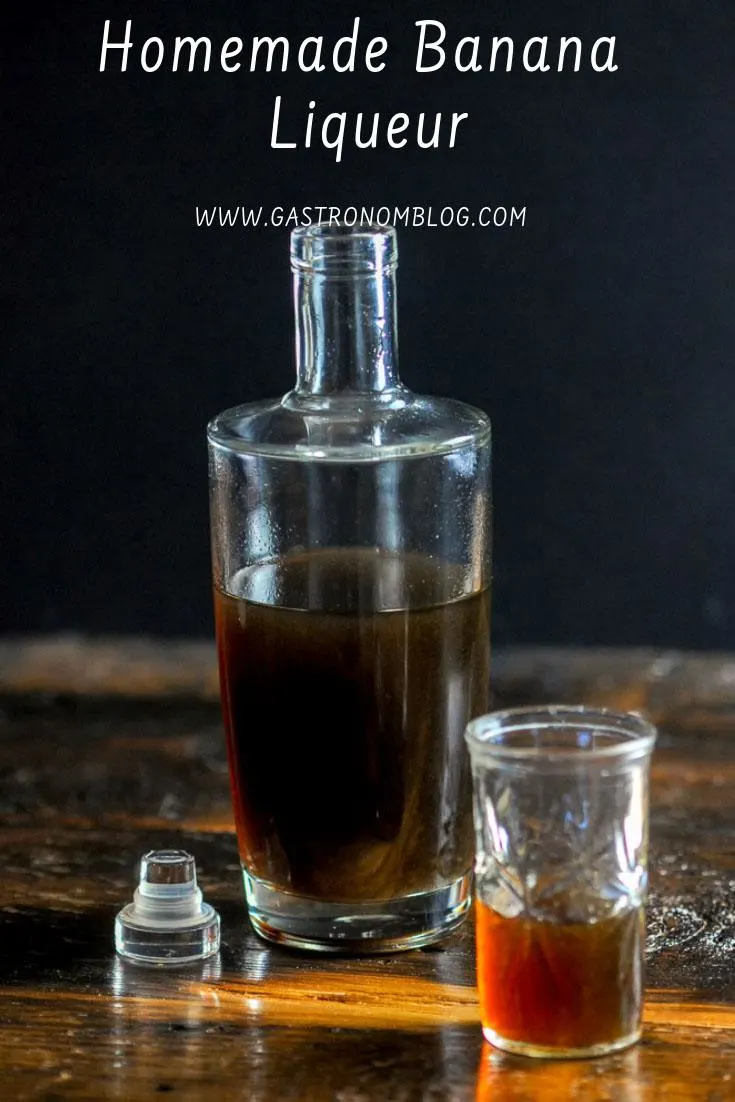 A bottle full of homemade banana liqueur sits on a wooden bar top next to a small shot glass with banana liqueur in it.