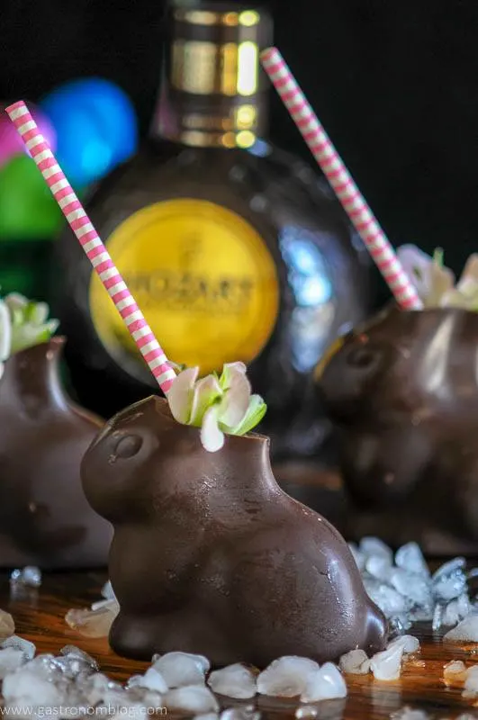 Chocolate Easter Bunnies filled with an Easter Bunny cocktail sit on a bar top garnished with spring flowers in front of a bottle of Mozart Dark Chocolate Liqueur.