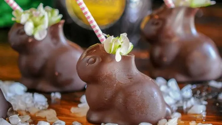 Chocolate Easter Bunnies filled with an Easter Bunny cocktail sit on a bar top garnished with spring flowers in front of a bottle of Mozart Dark Chocolate Liqueur.