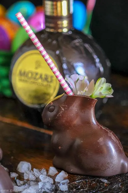 Chocolate Easter Bunnies filled with a cocktail sit on a bar top garnished with spring flowers in front of a bottle of Mozart Dark Chocolate Liqueur.