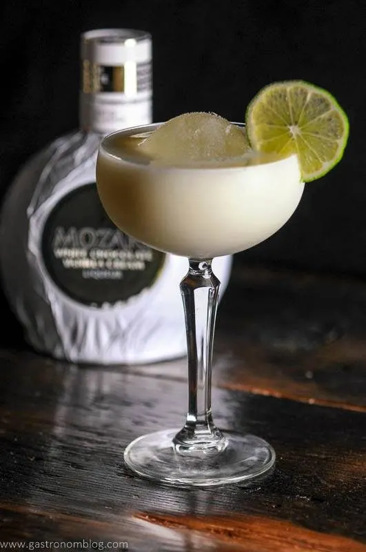 White Chocolate Martini in a coupe glass with ice ball, lime wheel, limes and bottle in background