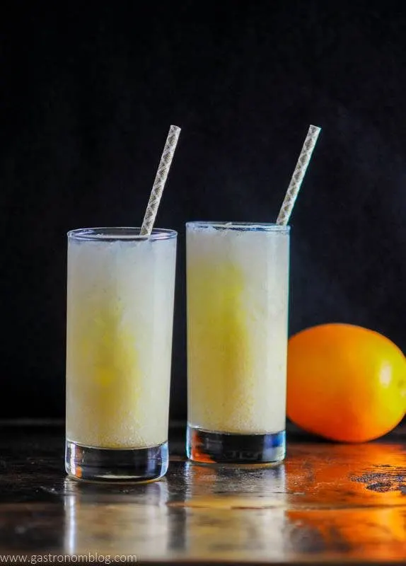 2 glasses with orange cocktail, purple straws and an orange in background