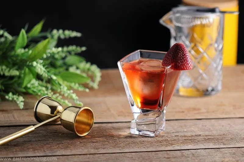 red cocktail with strawberry, mixing glass, jigger and flowers in background