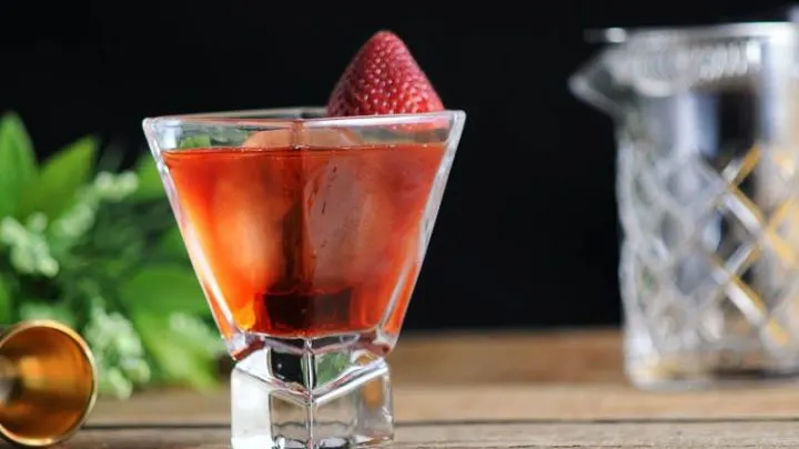 Pink cocktail in square glass with strawberry on rim. Mixing glass and greenery in background
