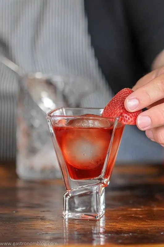 red cocktail in glass, strawberry being placed by a hand