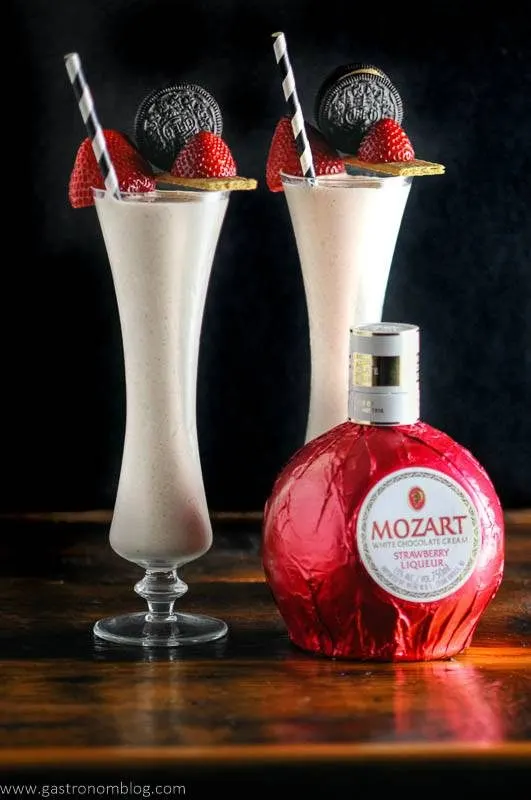 Strawberry milkshakes in tall glasses with oreos, strawberries, black and white straws with strawberry liqueur bottle. 