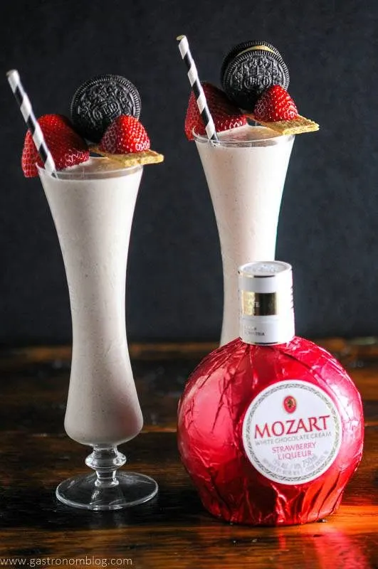 Strawberry milkshakes with strawberry liqueur bottle in front. Straws, strawberries, oreos and graham cracker garnishes. 