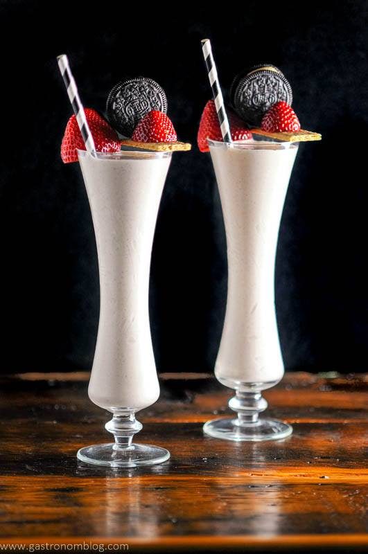 Strawberry milkshakes in tall glasses with black and white straws, strawberries, graham crackers and oreos as garnishes. 