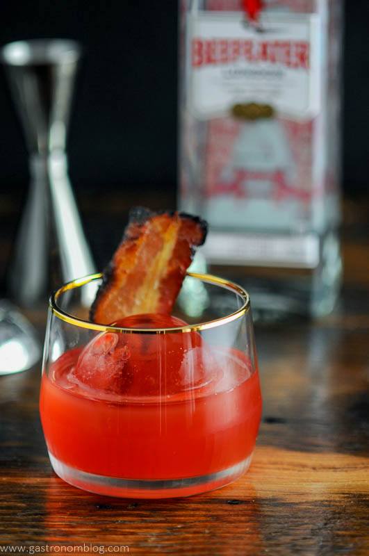 red Cocktail in gold rimmed rocks glass with bacon, beefeater bottle behind