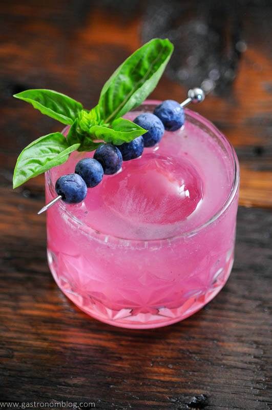 Top shot of pink cocktail, blueberries on cocktail pick, sprig of basil