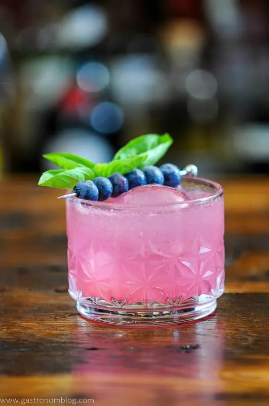 A pink cocktail in rocks glass with blueberries and basil as a garnish, this Blueberry Lemonade cocktail is on a wooden table.