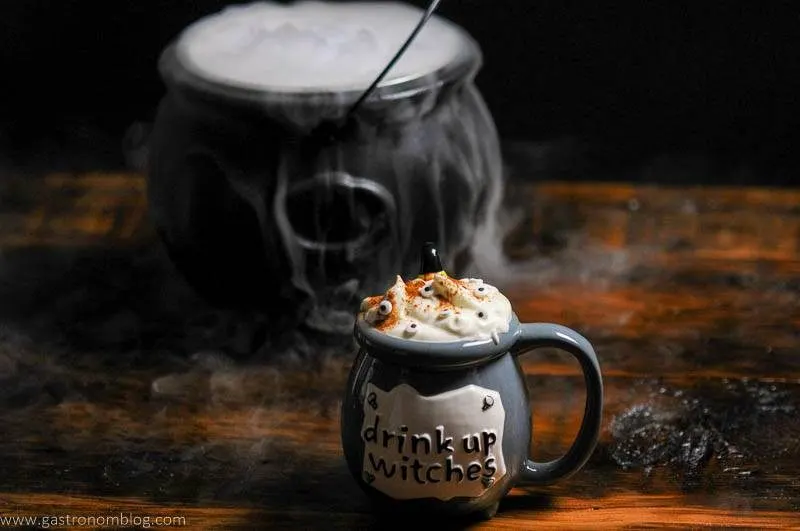 Witch Cauldron Halloween Cocktail, topped with whipped cream and eye candies. Cauldron with dry ice smog in back