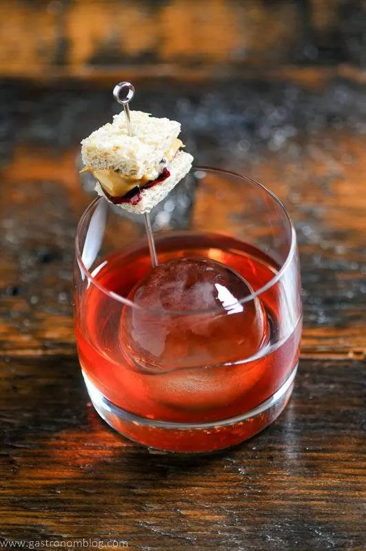 red colored Peanut Butter and Jelly Cocktail in rocks glass, large ice ball, tiny PB & J sandwich garnish
