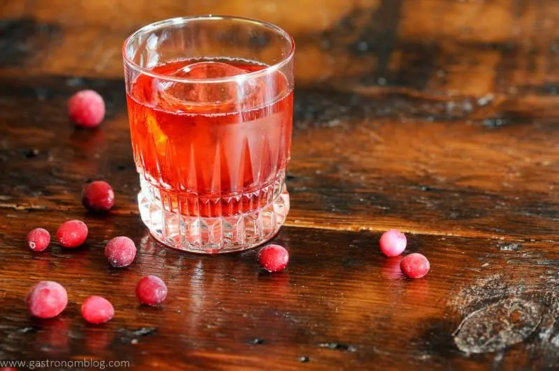red Negroni cocktail recipe in rocks glass, cranberries around glass on wood table