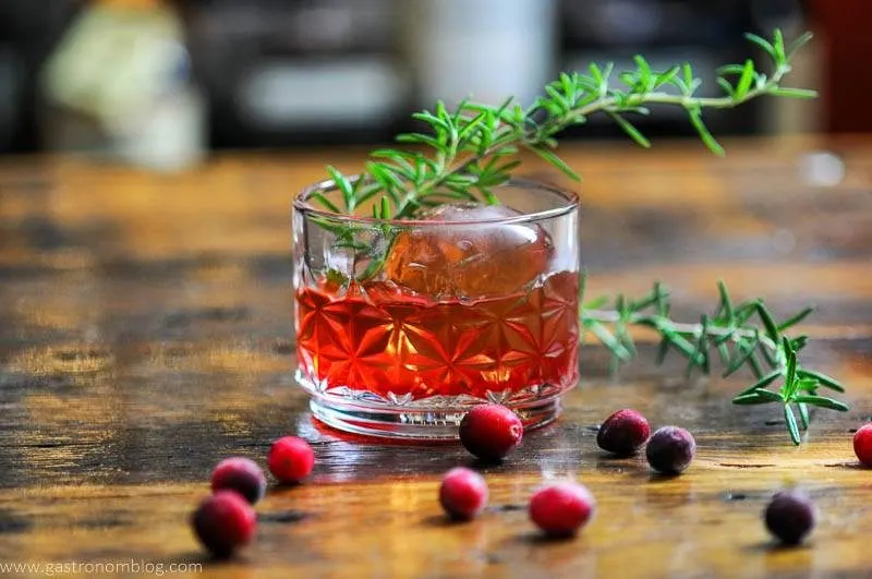 Red cocktail in rocks glass, rosemary sprigs, cranberries on wood table