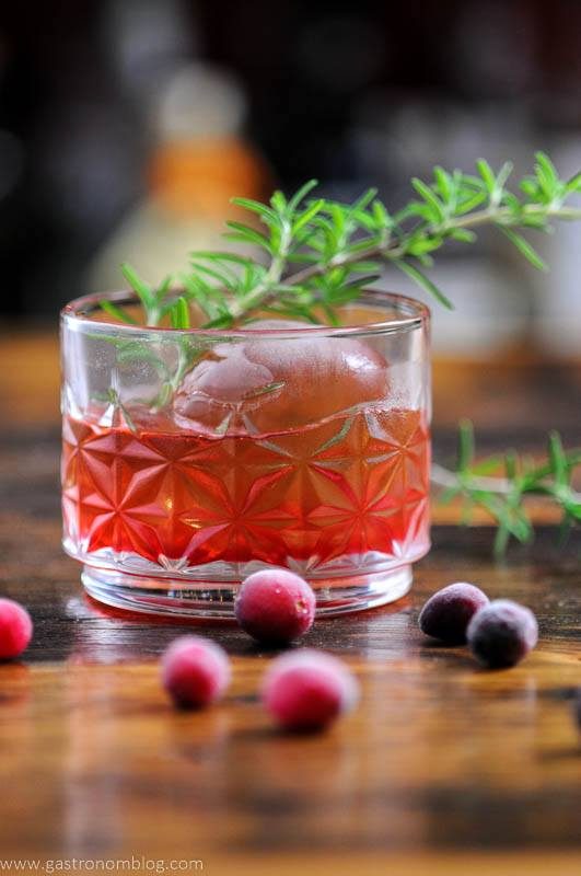 Cranberries around red cocktail in glass with rosemary sprigs