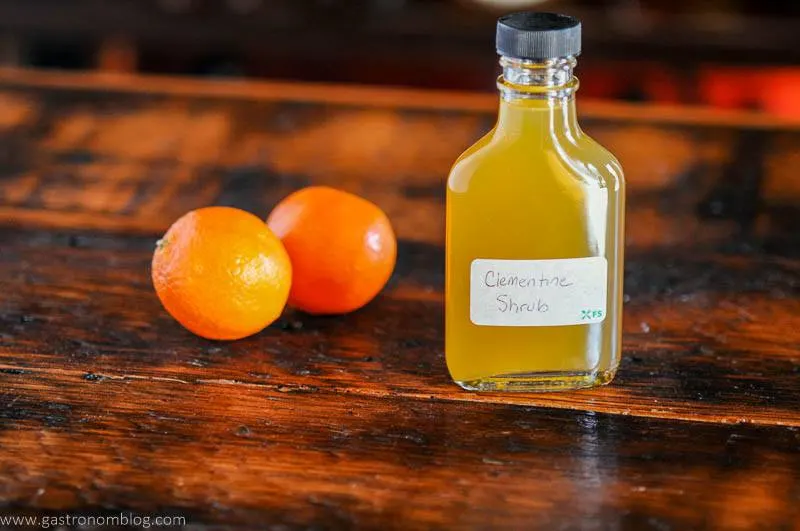 Orange shrub, or drinking vinegar recipe in a bottle with white label. 2 oranges behind on wooden surface