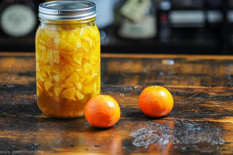 Orange shrub liquid in mason jar with 2 oranges on wooden surface.
