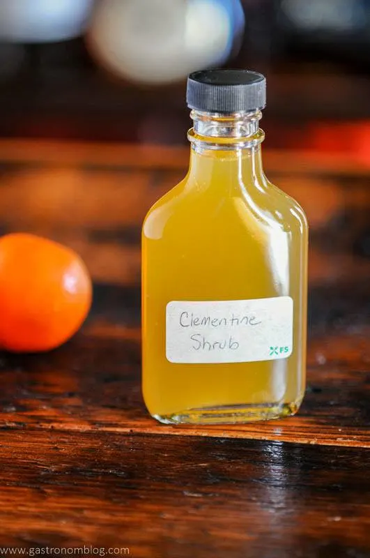 This orange drinking vinegar recipe is in a bottle with white label and black cap. orange behind on wooden surface