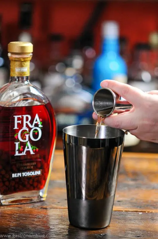 Liquor being poured from jigger into shaker tin, bottle behind