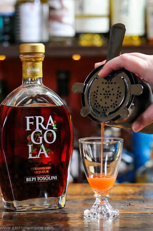 Strawberry cocktail being poured into a glass, bottle behind