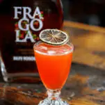 Pink cocktail in glass with citrus slice garnish, bottle behind