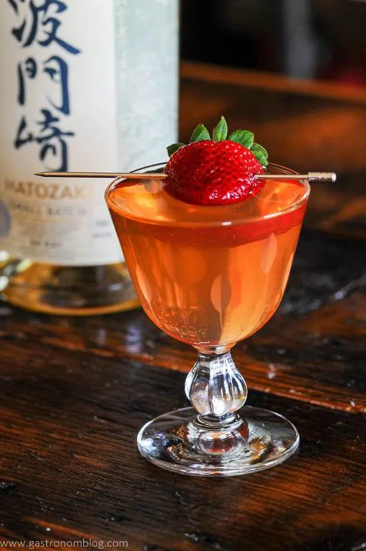 Pink cocktail in glass with strawberry on pick, whisky bottle in backgroun