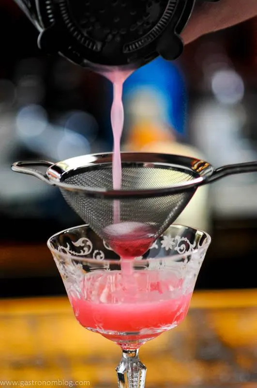 Pouring pink drink from shaker through mesh strainer into coupe