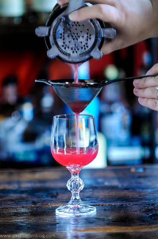 Hot pink cocktail being double strained with 2 strainers into cocktail glass