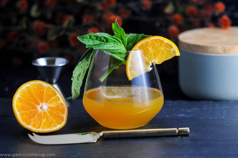 Tangerine drink in glass with orange slice and basil in glass. Sliced orange with knife and jigger next to glass. Gray container and orange flowers in background