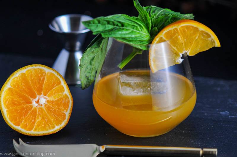 Tangerine drink in glass with orange slice and basil bunch. Sliced orange and silver knife and jigger next to glass.