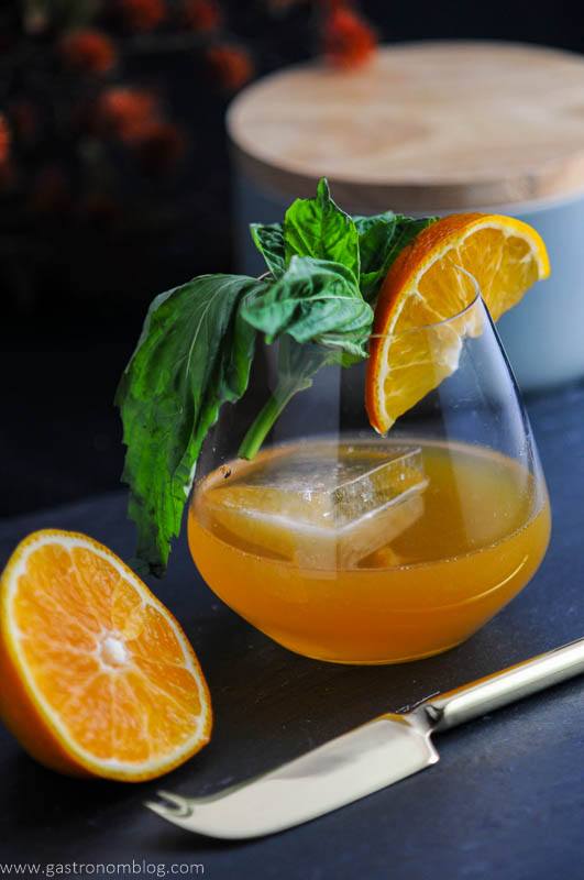 Orange cocktail in glass, this tangerine drink is with orange slice and basil. Orange slice and knife on slate