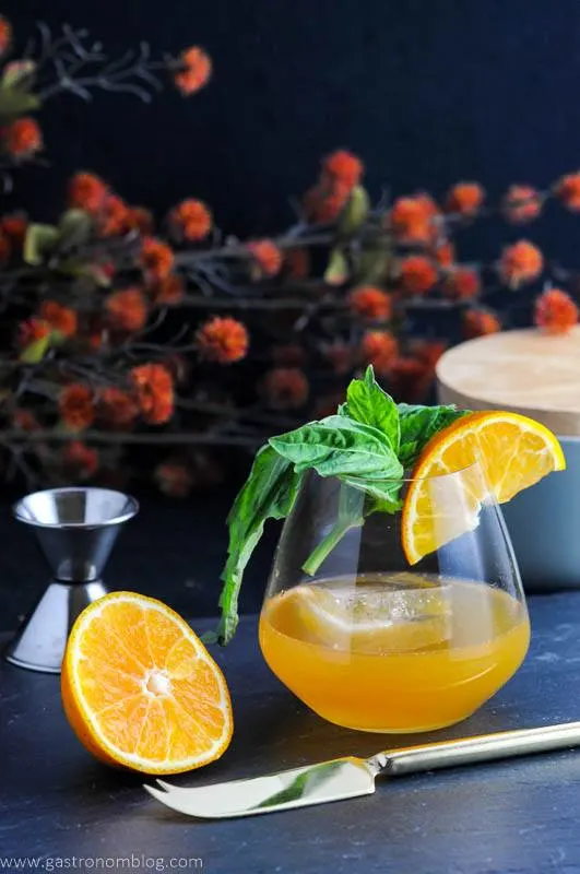 Tangerine drink in glass with basil and sliced orange. Orange sliced, silver knife and jigger next to glass. orange flowers behind