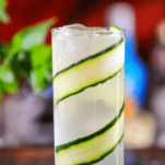 Cocktail in tall glass, cucumber slices line up sides, mint behind