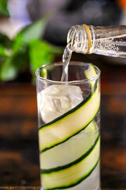 Tonic water being poured into tall glass with cucumber slices and clear ice cubes