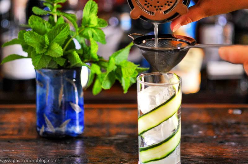 Cocktail being double strained into tall glassl ined with cucumber slices, blue glass with mint behind