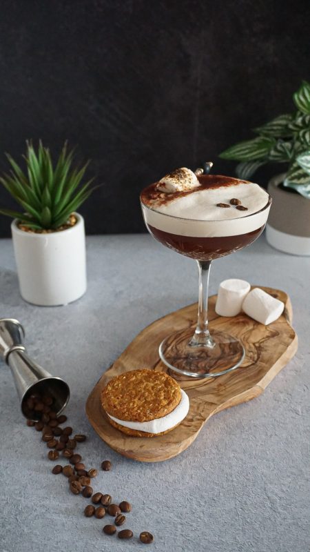Brown cocktail with white foam on wood board, marshmallows, jigger and plant in background