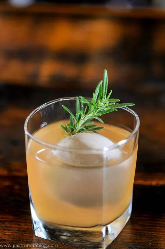 light orange cocktail in glass with ice and rosemary