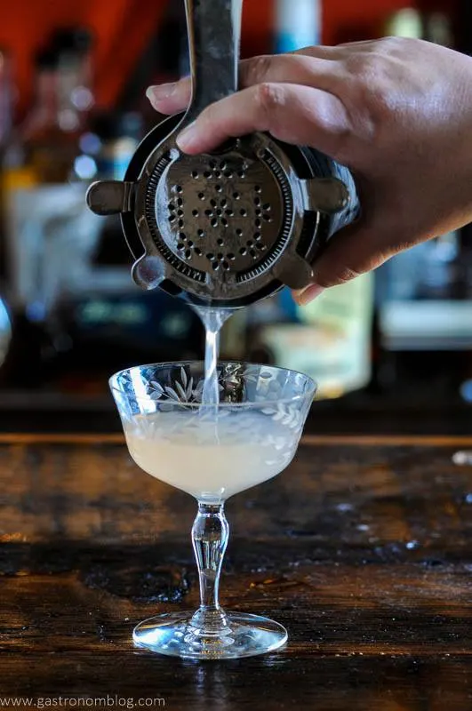cocktail being poured into a coupe glass from a shaker with a hawthorne strainer