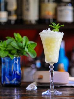 Absinthe Frappe in tall glass with crushed ice and mint garnish, mint in blue vase behind