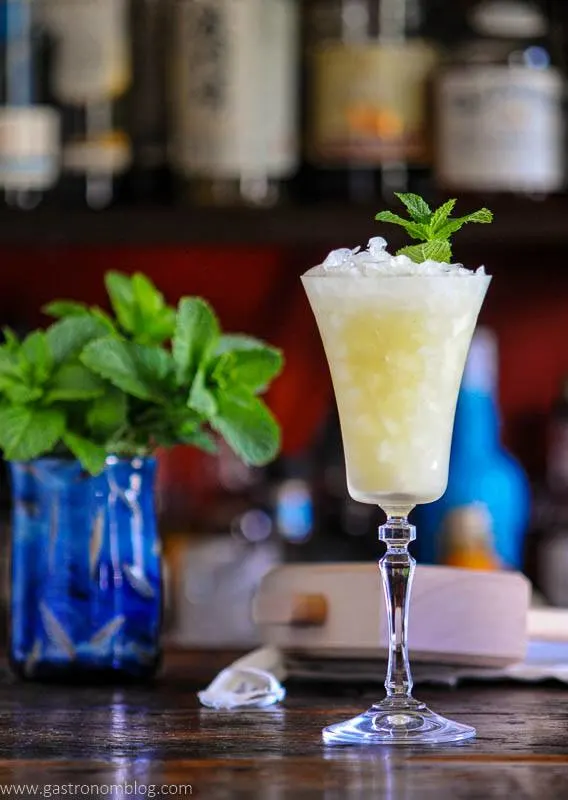 Absinthe Frappe in tall glass with crushed ice and mint garnish, mint in blue vase behind