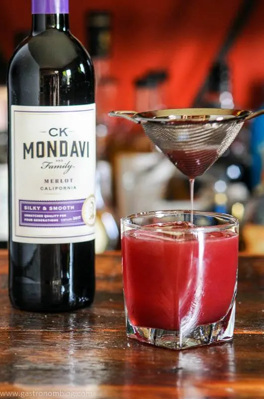 Red cocktail being poured into glass, red wine bottle behind