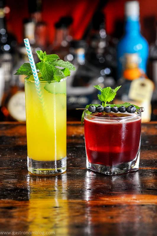 2 cocktails on wood board - orange cocktail in tall glass, red cocktail in rocks glass