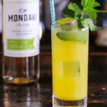 Yellow cocktail with mint in tall glass with straw, white wine bottle behind