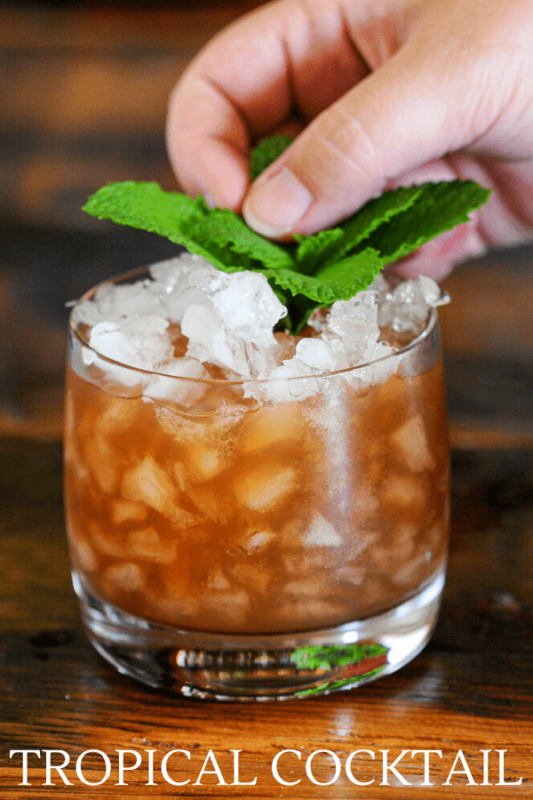 Orange cocktail in rocks glass with crushed ice, mint sprig being placed by hand