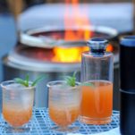 Orange cocktail in 2 glasses and decanter on blue table, fire behind
