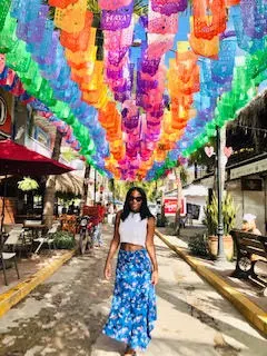 Lady in white top, blue skirt walks under rainbow colored lanterns in alleyway