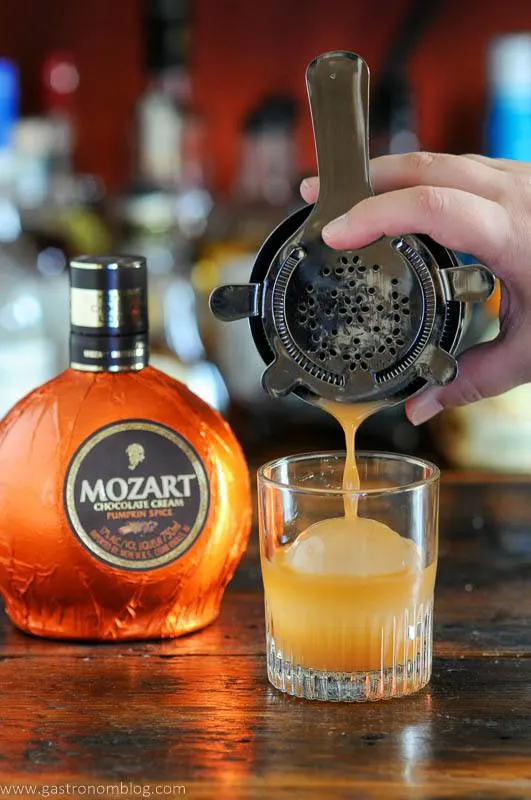 Orange Pumpkin cocktail being poured into a rocks glasses with clear ice ball, orange liqueur bottle behind