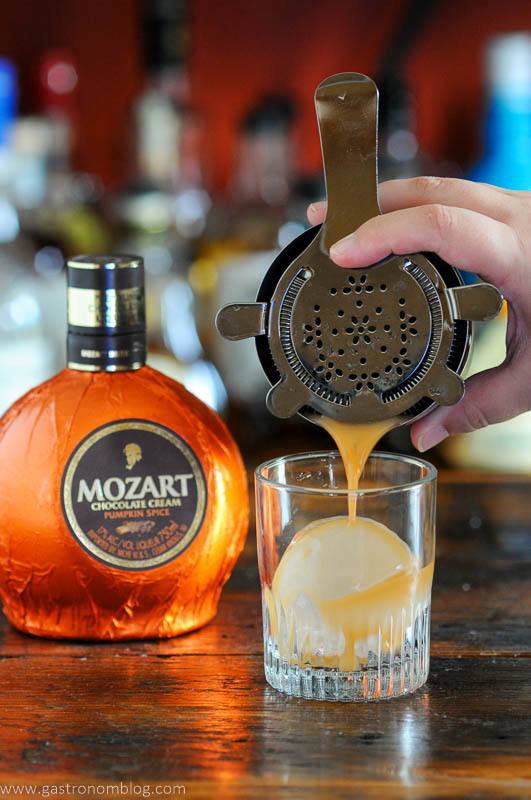 Orange Pumpkin cocktail being poured into a rocks glasses with clear ice ball, orange liqueur bottle behind