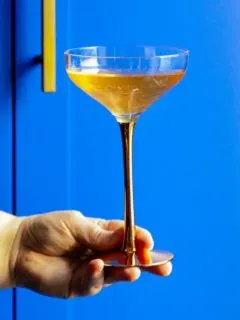 Bridgerton cocktail, a golden color in a copper stemmed coupe against a blue background held by a hand